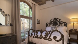 Een slaapkamer met een groot raam, een bed met een decoratief hoofdeinde, een nachtkastje met een lamp en een dressoir.
