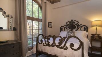 Un dormitorio con un ventanal grande, una cama con cabecero decorado, una mesita de noche con lámpara y un aparador.