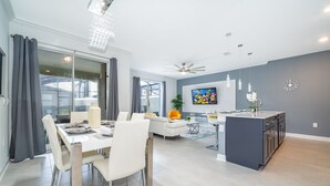 Une cuisine moderne dotée d’un îlot central, un coin repas avec une table et des chaises blanches, et un espace de détente avec un canapé et un téléviseur.