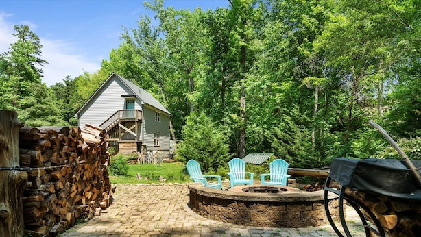 Un chalet rustique doté d’un foyer en pierre, entouré d’une zone boisée et d’une pile de bois de chauffage.