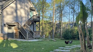 Une maison avec un escalier en bois menant à un porche, entourée d’une cour verdoyante et d’arbres.