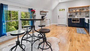 Une cuisine moderne avec des meubles de couleur sombre, un plan de travail blanc et un four encastré. On y trouve un coin repas avec une table ronde et des tabourets, ainsi qu’une grande fenêtre ornée de rideaux bleus.