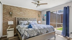 A bedroom with a brick wall, a bed with a grey and white patterned bedspread, a ceiling fan, and a window with blue curtains. - 5 STAR Townhome near all major highways! Fast Wifi (Carrollton)
