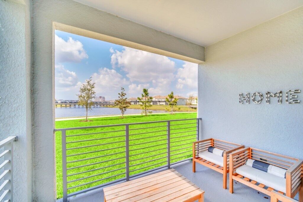 A balcony with a view of a green park and a body of water, two wooden lounge chairs, and a table.