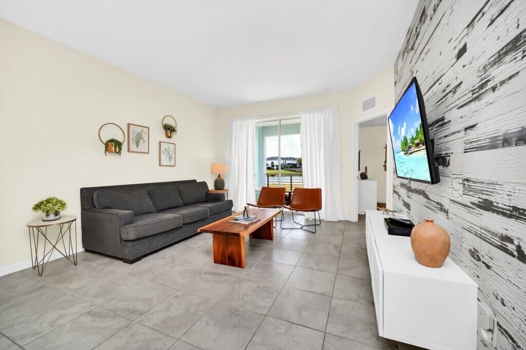 Un salon moderne avec un canapé gris, une table basse en bois et un téléviseur fixé à un mur blanc. On y trouve deux fauteuils et une petite table d’appoint avec une plante.