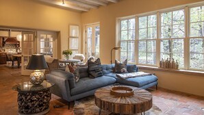 A cozy living room with a sectional sofa, a round coffee table, and a large window with a view of trees. - Tranquil compound directly across from 10,000 waves and close to town and skiing (Santa Fe)