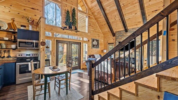 A spacious cabin interior with a kitchen, dining area, and a staircase leading to a loft bedroom. - EagleRock Retreat | Pet Friendly w/ Hot Tub, Firepit + WiFi (Rogers)