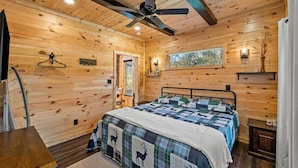 A cozy log cabin bedroom with a bed, wooden walls, a ceiling fan, and a window with a view of trees. - EagleRock Retreat | Pet Friendly w/ Hot Tub, Firepit + WiFi (Rogers)