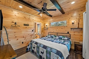 A wooden cabin bedroom with a bed, a ceiling fan, a window, and a door. - EagleRock Retreat- Pet Friendly, Hot Tub, Firepit, WiFi (Rogers)