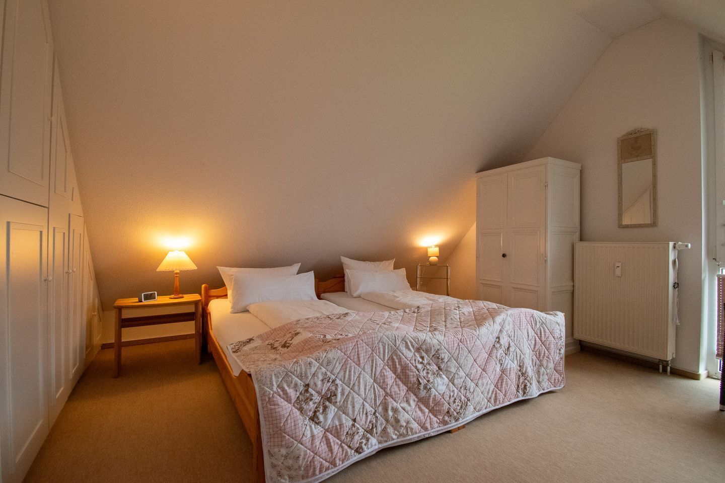 A bedroom with a large bed, two bedside tables with lamps, a wardrobe, and a radiator.