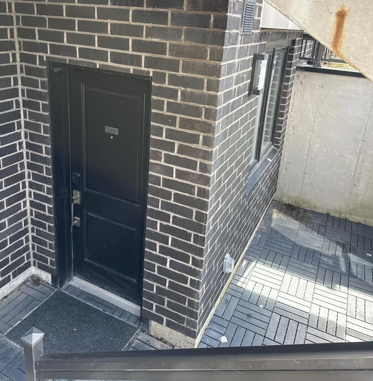A black door with a number plate, a brick wall, and a tiled ground.