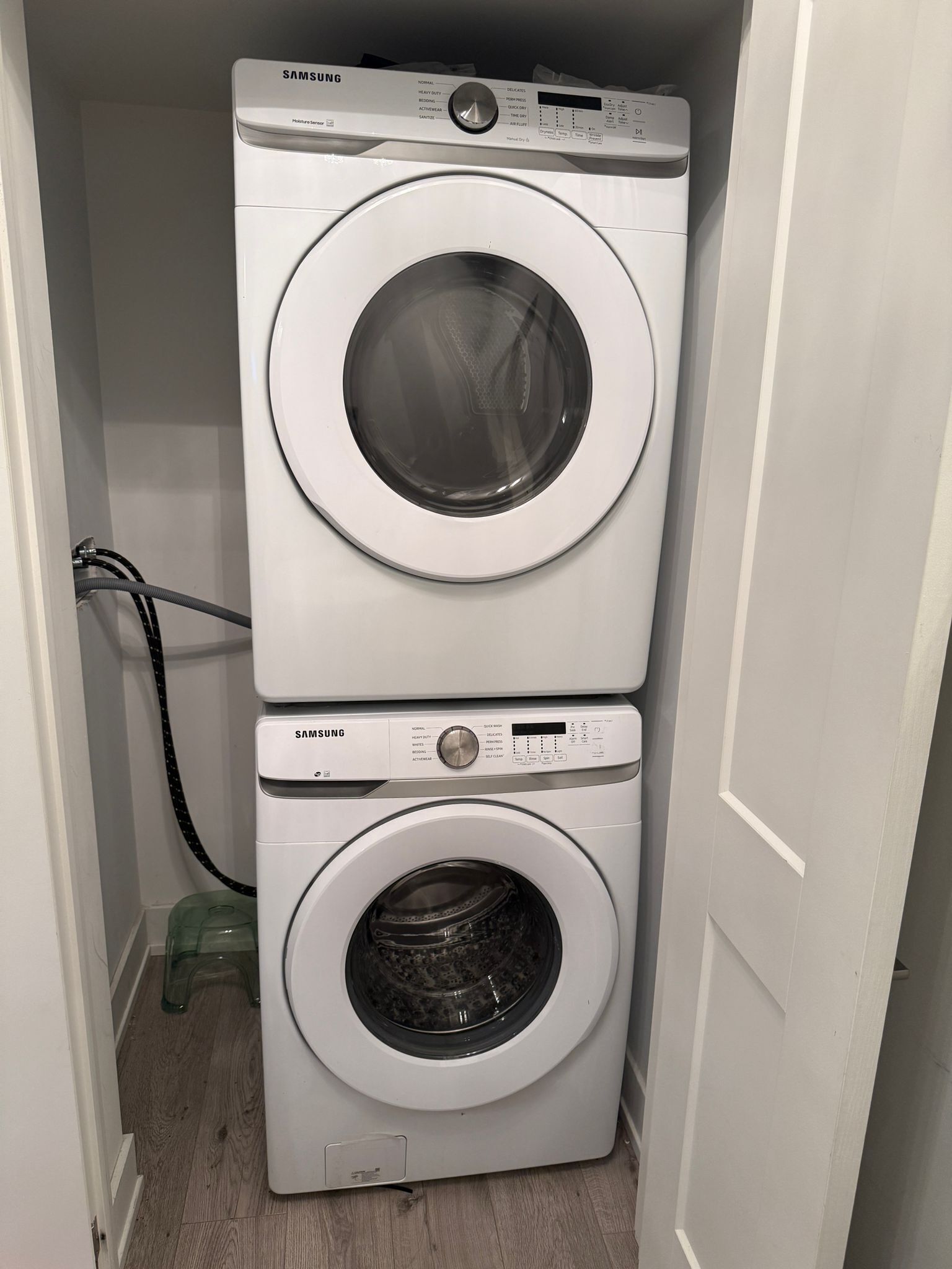 A stacked washer and dryer unit in a small laundry room with a visible power cord and a green trash bin.