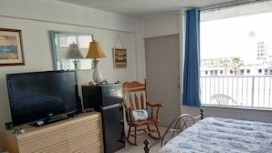 A hotel room with a bed, a desk with a TV, a refrigerator, a chair, and a window with a view of buildings.