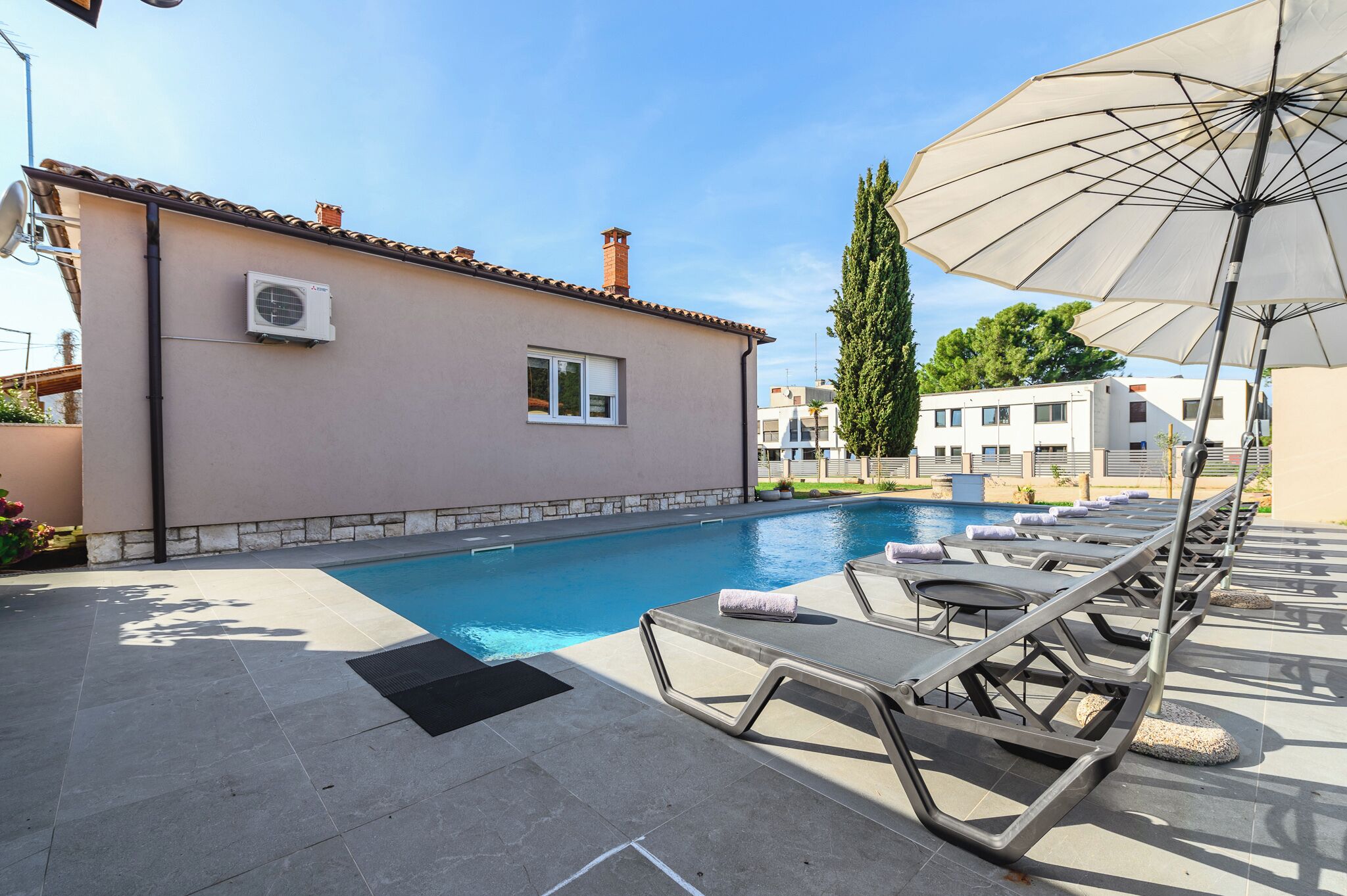 A swimming pool with lounge chairs and an umbrella, a building with a chimney, and a clear sky.