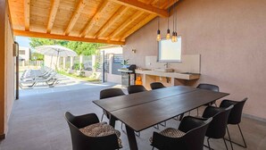 A covered outdoor dining area with a long table and chairs, a grill station, and a view of a garden with sun loungers. - Villa with pool, air conditioning, internet and covered terrace, near the sea (Umag)
