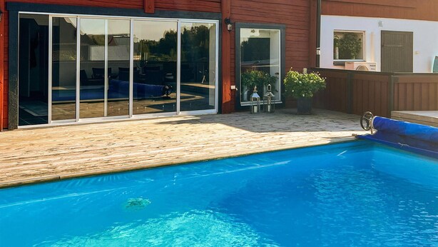 En pool med en trädeck, skjutbara glasdörrar och en liten uteplats med utemöbler.