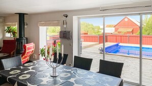 En täckt uteplats med ett matbord, stolar, en svart spis och utsikt över en swimmingpool och ett rött staket.