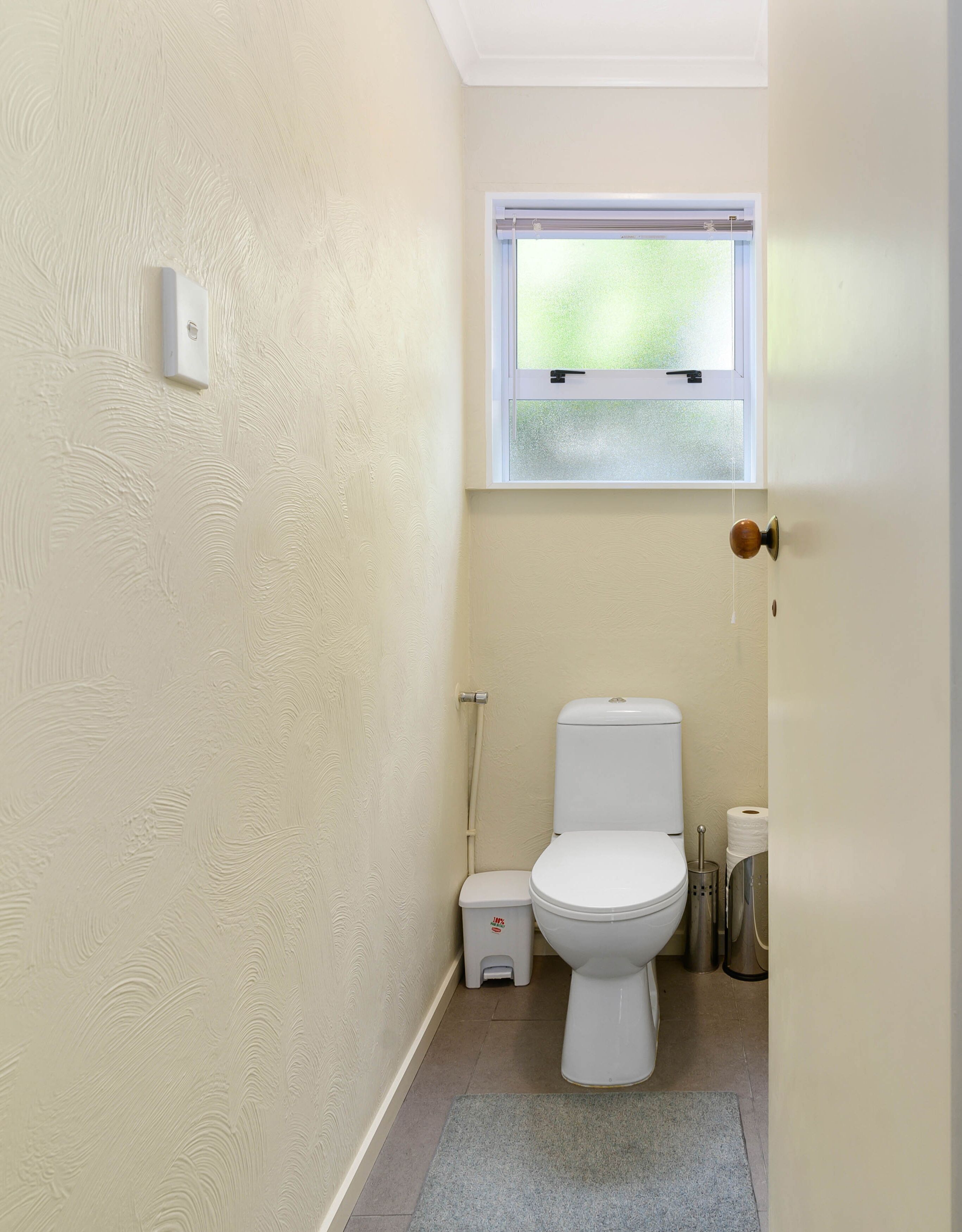 A compact bathroom with a toilet, a small trash bin, and a window above the toilet.