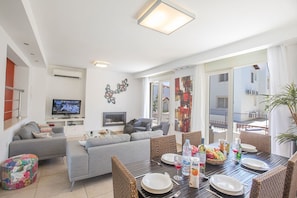 A modern living room with a grey sofa, a dining table set for four, and a television in the background.