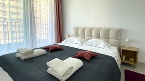 Een net opgemaakt bed met een donge deken, witte kussens en twee sierkussens. Er is een raam met dunne gordijnen en een uitzicht op gebouwen.