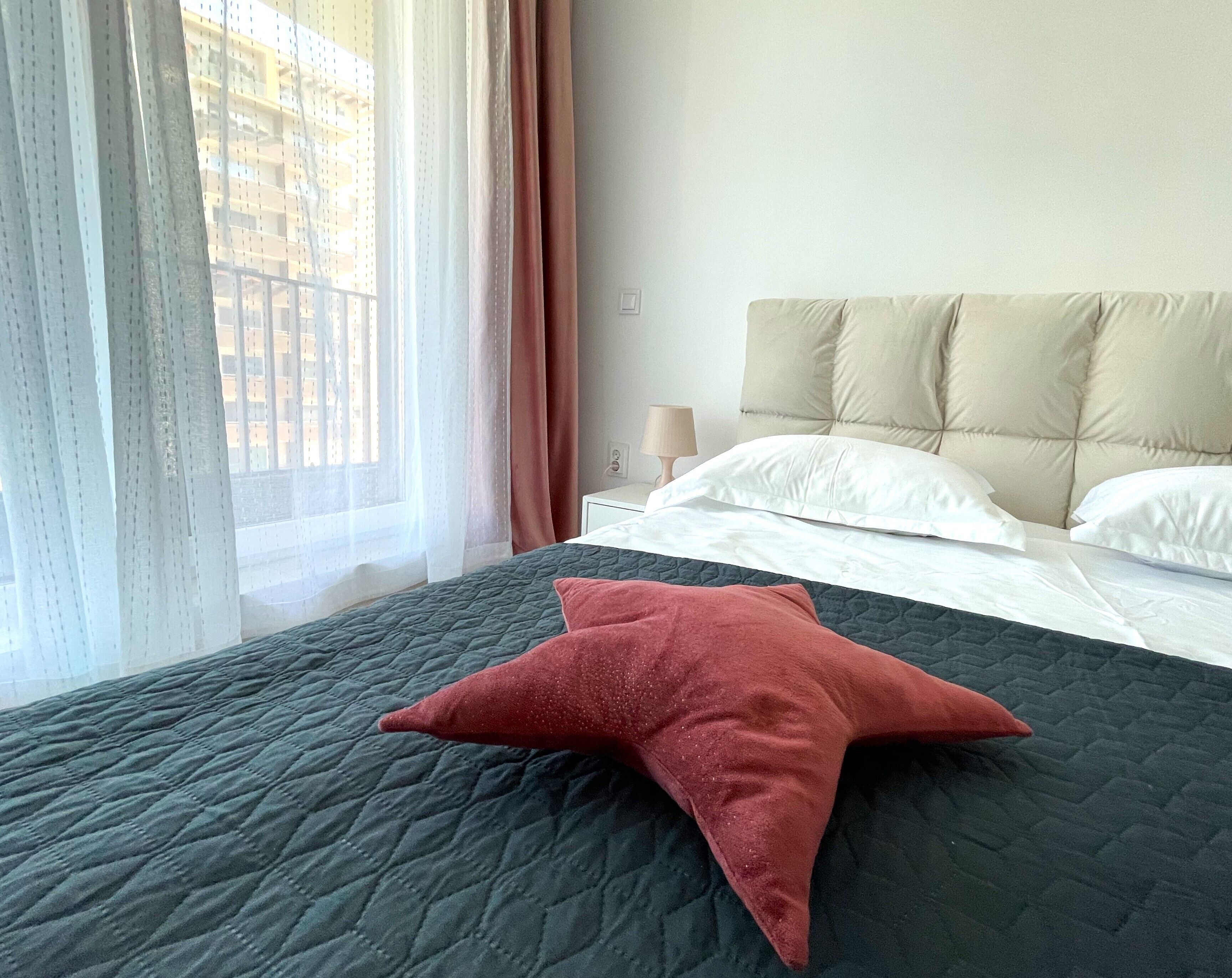 Een bed met een donkere quilt en een rood stervoormig kussen, een wit hoofdeinde en een raam met dunne gordijnen en uitzicht op gebouwen.