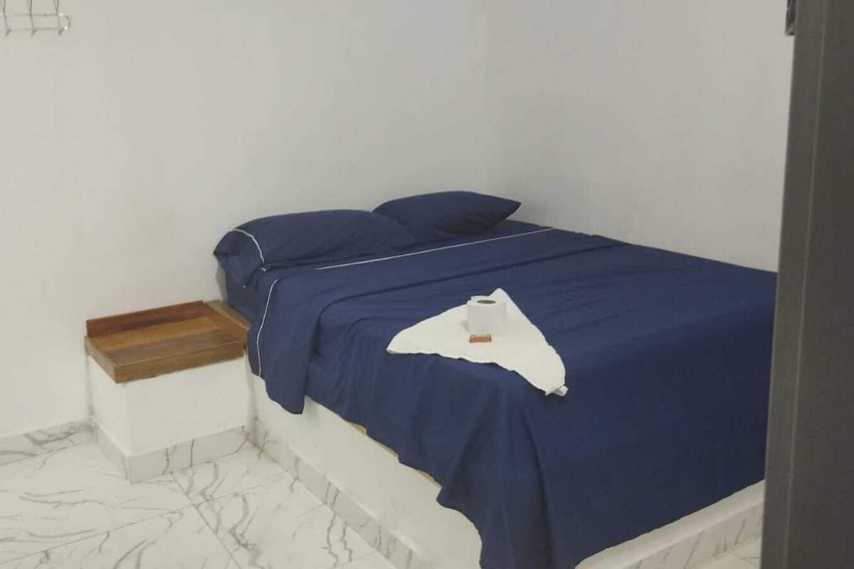 Uma cama pequena e bem arrumada, com cobertura e almofadas azuis, um criado-mudo de madeira com gaveta, e um chão com padrão marmorizado.