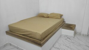 Una cama con colchón y almohadas color crema, una mesita de noche con tapa de madera y un suelo con estampado de mármol.