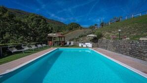 Una piscina con sillones de descanso y un pabellón, rodeada de muros de piedra y vegetación.
