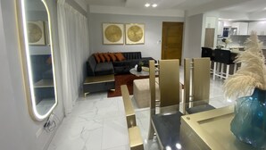 Un salon moderne avec un canapé, une table basse et des miroirs décoratifs. On y trouve également un coin repas avec des chaises et une table en verre. La pièce présente un design épuré et minimaliste, avec des murs blancs et un sol carrelé.