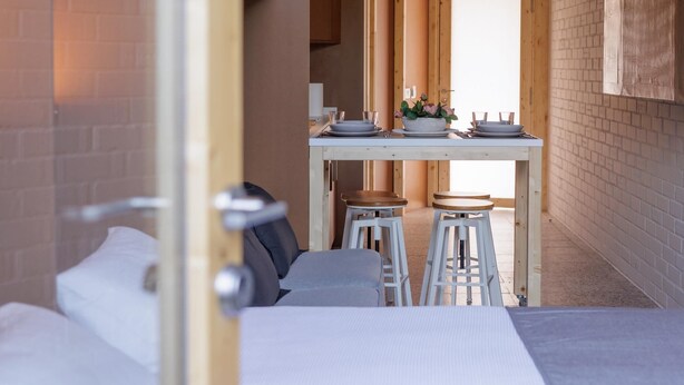 A room with a bed, a small table with stools, and a view of a brick wall and a door. - Apartamento estudio con terraza-pensión* (A Estrada)