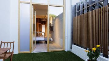 A modern, compact living space with a sliding glass door leading to a bedroom, a small patio area with a table and chairs, and a wooden fence.