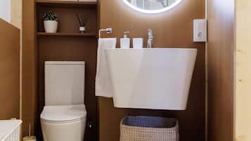 Une petite salle de bain avec des toilettes blanches, un lavabo surmonté d’un miroir et une petite étagère où sont posés des serviettes et un pot de fleurs.