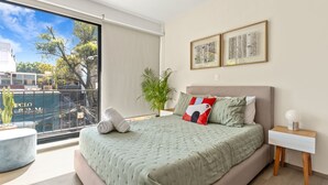 Une chambre à coucher moderne avec un grand lit, une table de chevet, une plante en pot et une vue sur l’extérieur.