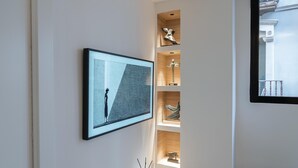 Un interior moderno con una obra enmarcadas, una consola blanca con libros y un jarrón, y una estantería de madera con objetos decorativos.