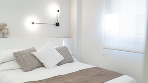 Una cama bien hecha con una manta blanca y marrón, dos almohadas y una lámpara de pared.