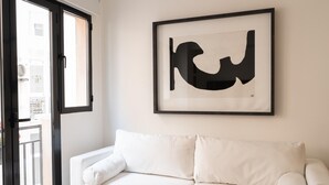 Un sofá blanco con una obra de arte en un marco negro en la pared, una puerta corredera de vidrio y vistas a edificios.
