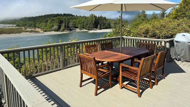 Une terrasse en bois avec une table et des chaises, un barbecue et une vue sur un lac et une forêt.