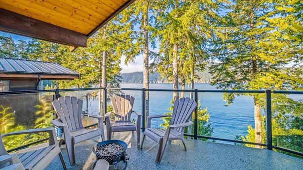 Eine überdachte Terrasse mit Adirondack-Stühlen und einem Feuerkorb, mit Blick auf einen See und Wald.