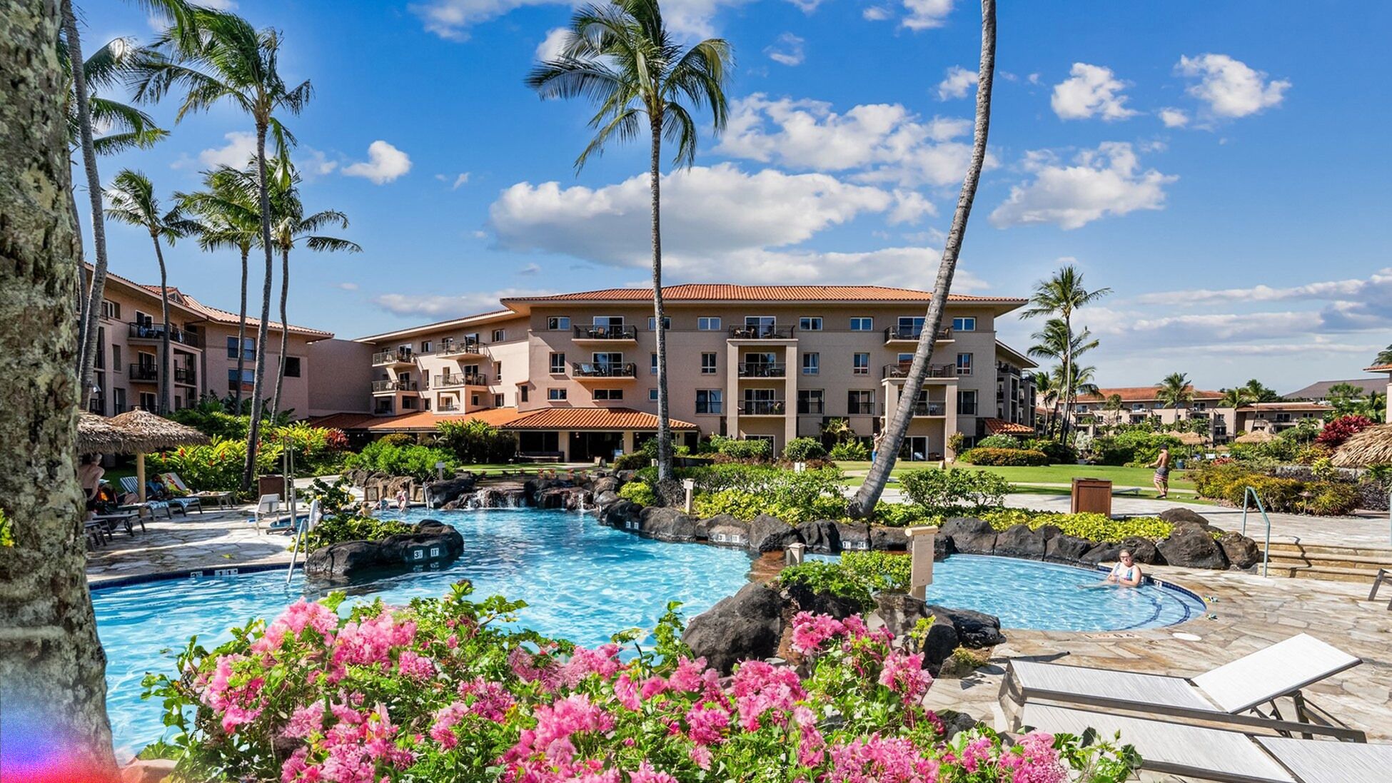 A resort with a central swimming pool, palm trees, and a multi-story building with balconies.