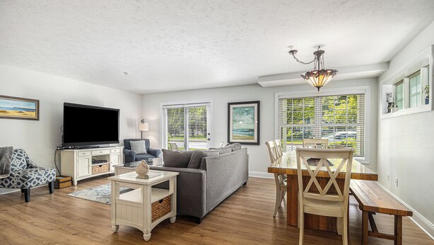 A living room with a grey sofa, a TV, a dining table with chairs, and large windows with blinds.