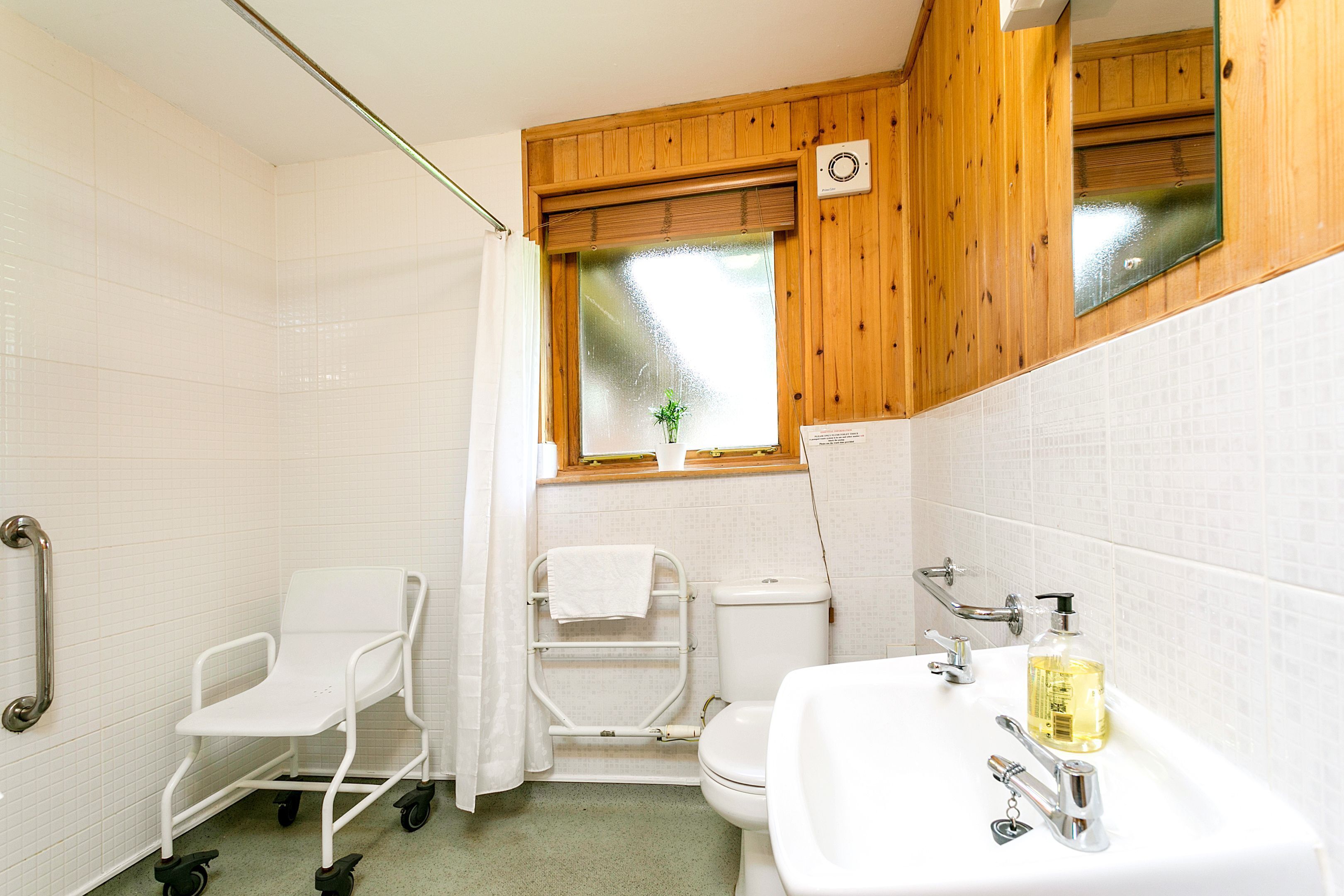 A bathroom with a white sink, toilet, and shower area, wooden walls, and a window with a plant.