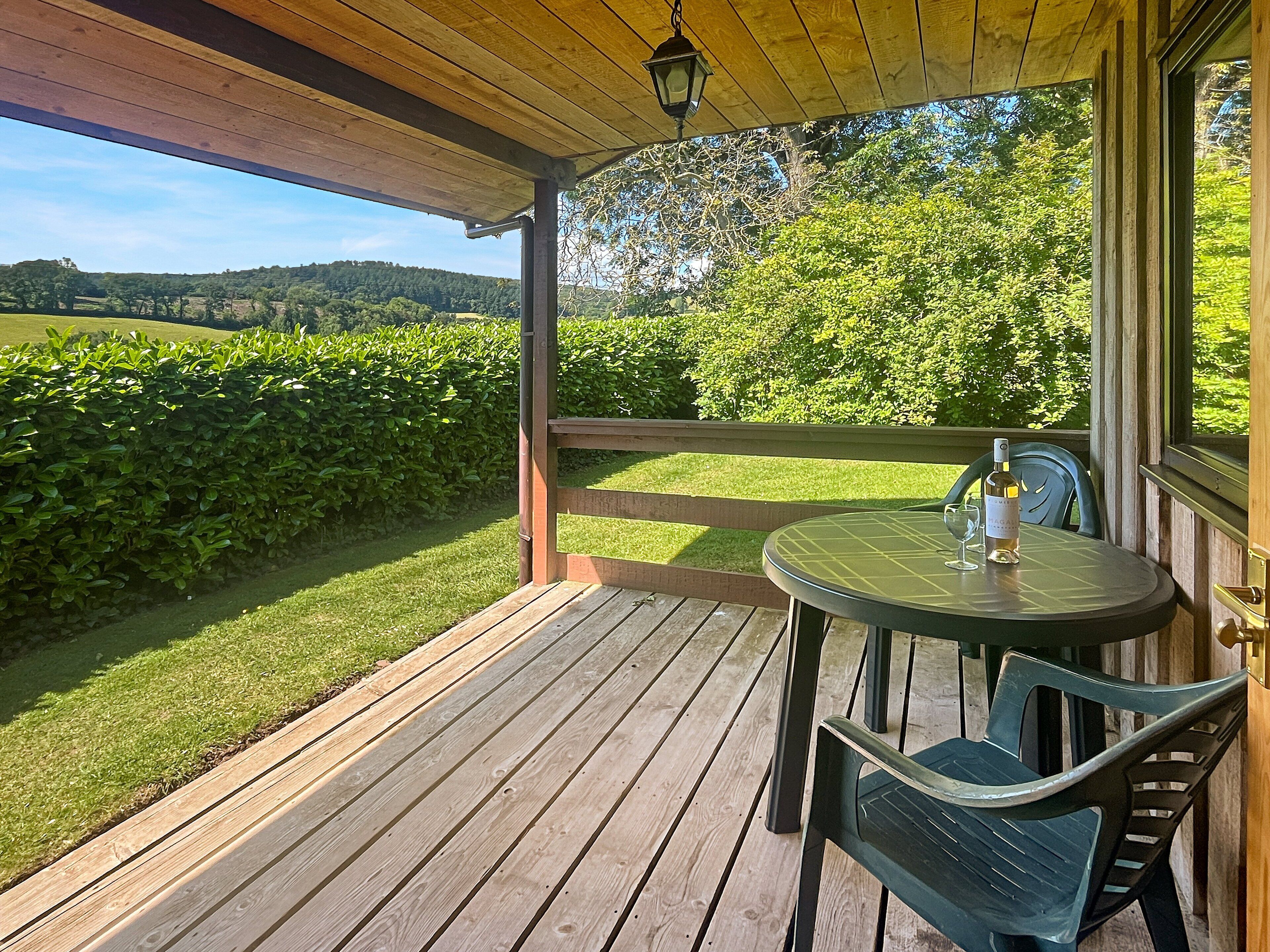 A wooden deck with a table and chairs, overlooking a green landscape with trees and hills.