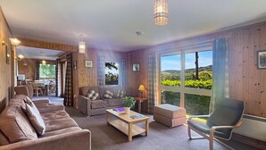 Ein gemütliches Wohnzimmer mit einer Couch, Sesseln, einem Couchtisch und einem Essbereich mit Tisch und Stühlen. Große Fenster bieten Blick ins Grüne.