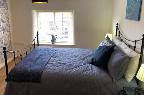 Une chambre à coucher avec un lit, une fenêtre avec des stores, une lampe et un vase posé sur le rebord de la fenêtre.