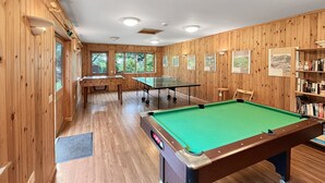Una taula de bitlles, una taula de billar i una taula de ping-pong en una sala amb panells de fusta, amb prestatgeries i quadres a les parets.