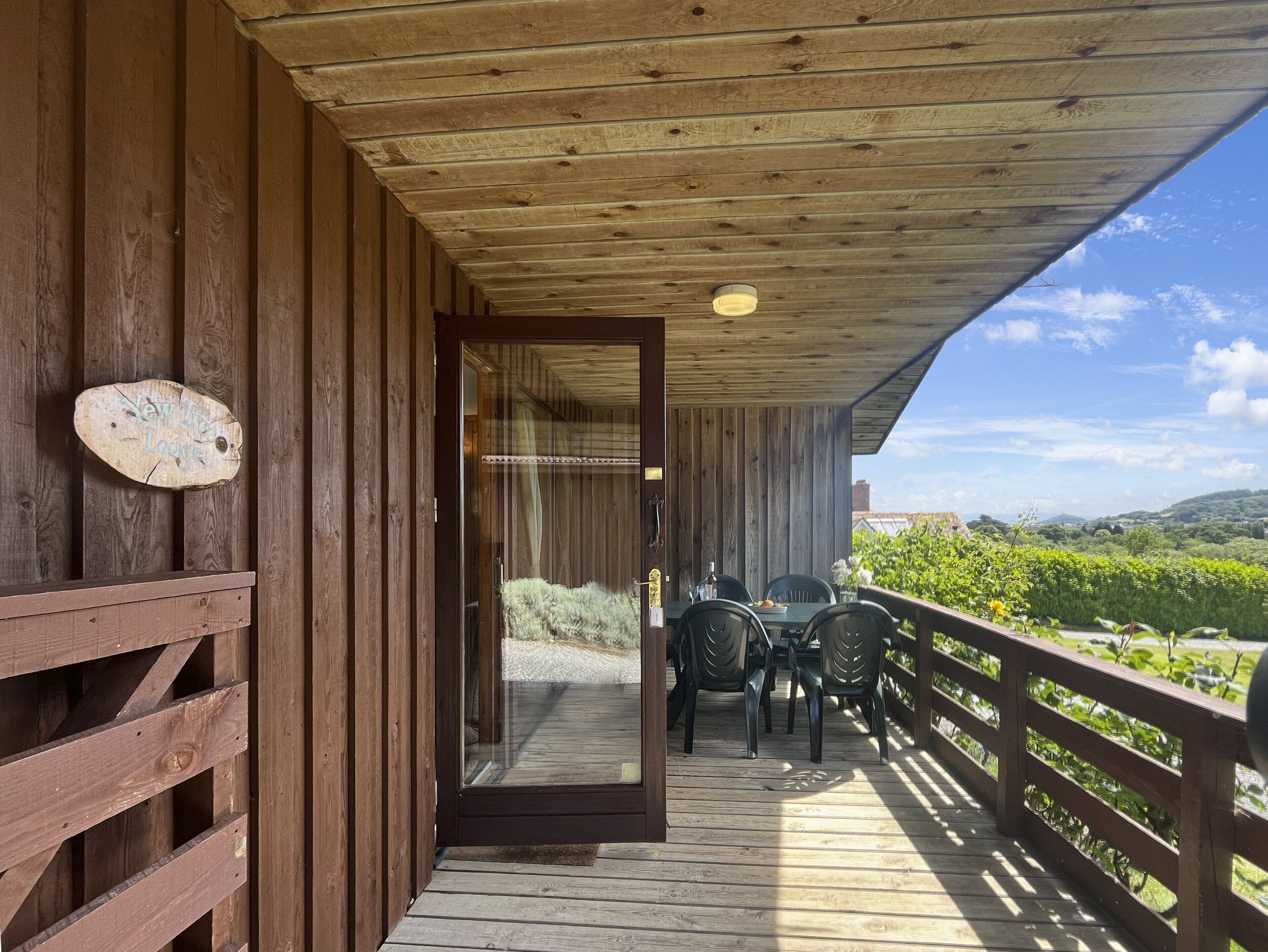 A wooden deck with a table and chairs, a wooden railing, and a view of greenery and hills.