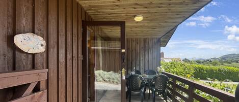 A wooden deck with a table and chairs, a wooden railing, and a view of greenery and hills.