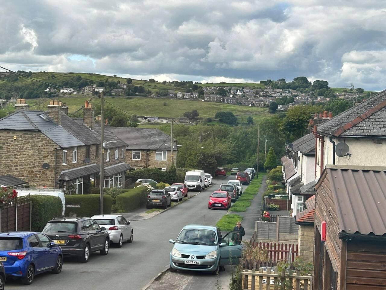 A residential street with houses on both sides, parked cars, and a hilly landscape in the background.