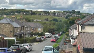 A residential street with houses on both sides, parked cars, and a hilly landscape in the background.