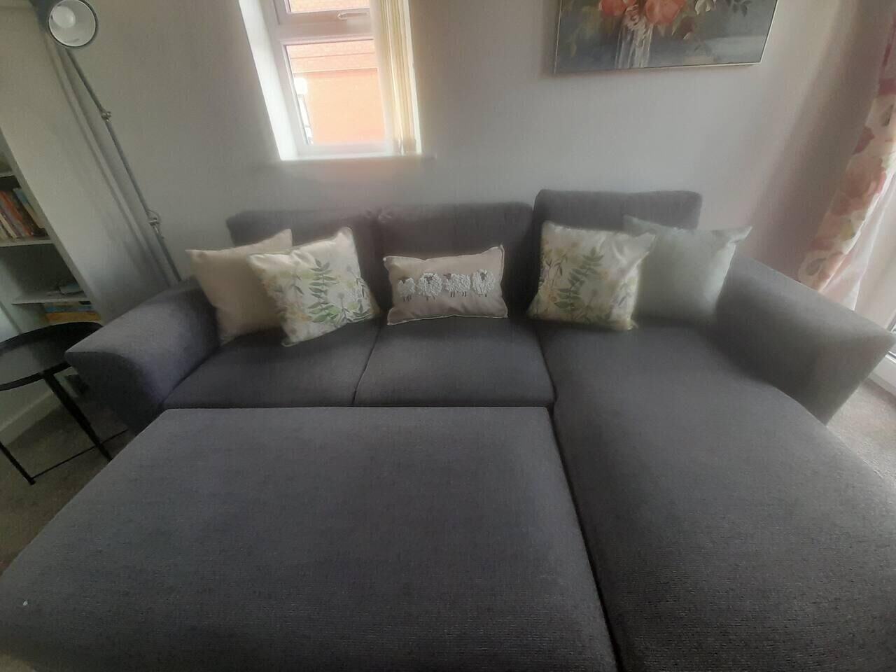 A grey sofa with floral cushions, a small side table, and a bookshelf in a room with a window and a painting on the wall.